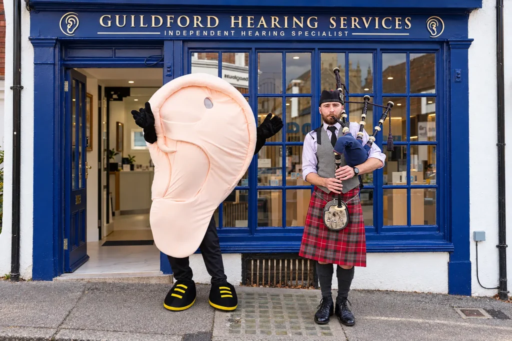 A bagpiper in Highland dress and a giant ear mascot outside the blue-fronted Guildford Hearing Services clinic