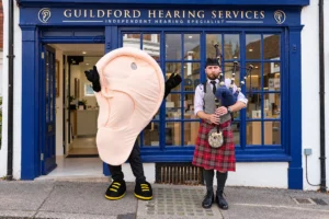 A bagpiper in Highland dress and a giant ear mascot outside the blue-fronted Guildford Hearing Services clinic